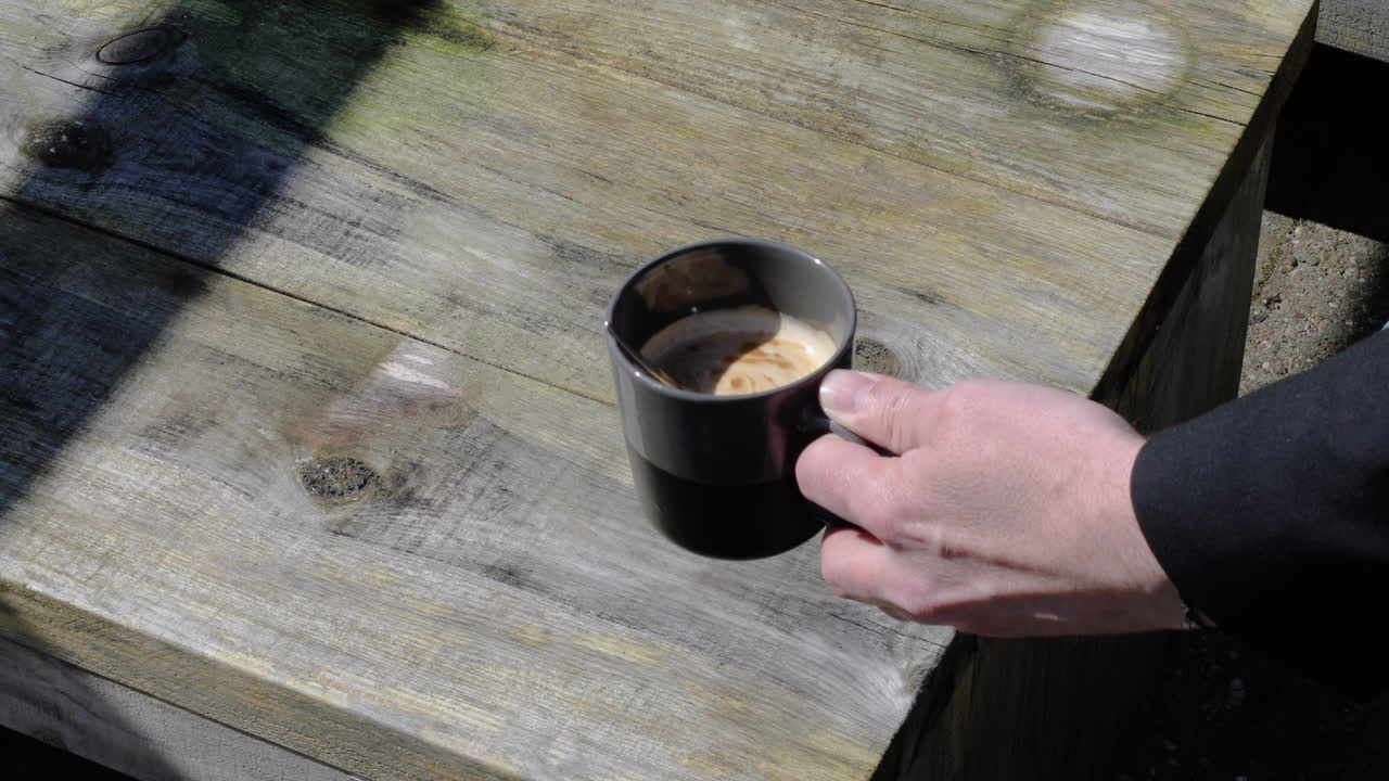 야외 나무 테이블의 배경 위에 검은 컵에 금속 스푼으로 커피를 휘젓는 남자 손의 클로즈업 샷