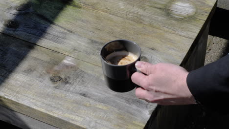 Primer-Plano-De-Una-Mano-De-Hombre-Revolviendo-El-Café-Con-Una-Cuchara-De-Metal-En-Una-Taza-Negra,-Sobre-Un-Fondo-De-Mesa-De-Madera-Al-Aire-Libre