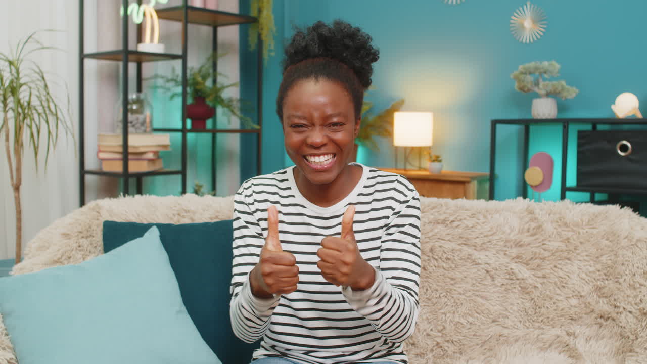 Donna afroamericana che celebra la vittoria, salta sul divano con le mani alzate, piena di gioia ed energia per il successo, in un ambiente interno.