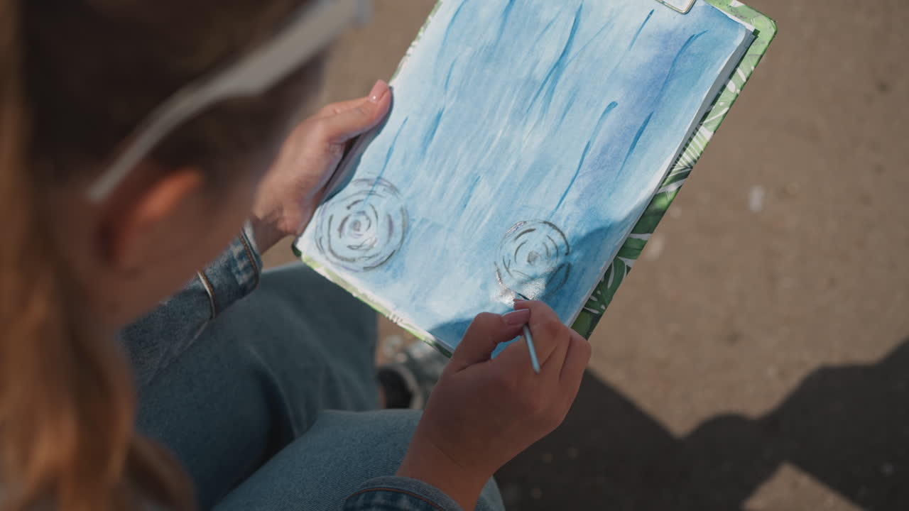 Artista ao ar livre criando pintura inspirada em música. Mulher engajada em expressão artística urbana com fones de ouvido. Artista feminina relaxada imersa em cena urbana ao ar livre pintando com inspiração musical.