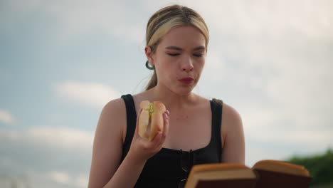 woman in black tank top eating burger while reading a book outdoors under bright sky, the book slightly blurred as she focuses on the food