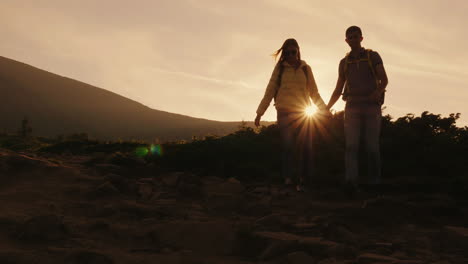 A-Young-Couple-Of-Tourists-With-Backpacks-Walking-Along-A-Mountain-Trail-In-The-Rays-Of-The-Setting-