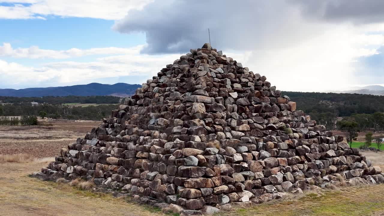 A stationary camera slowly zooms toward a large stone pyramid in a dry, open landscape. Daylight and passing clouds create dynamic natural lighting