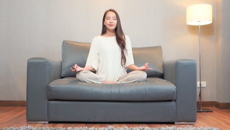 Yoga-and-meditation,-young-asian-woman-on-couch-in-easy-pose-aka-sukhasana-with-gyan-mudra-finger-gesture,-slow-motion