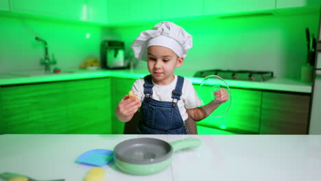 lovely baby chef in white cap stands at the table in the kitchen. baby boy opens toy frying pan and takes toy food.