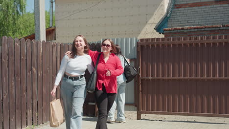 two ladies happily walk into compound through open gate under bright sky, one holds arm around neck of other as they step onto paved courtyard near wooden fence and green grass