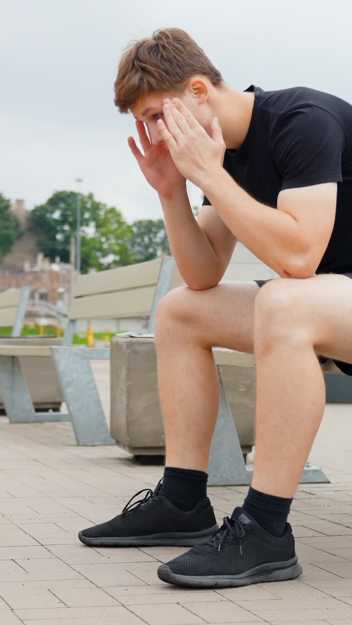 Vertical closw up of Young man sitting on a park bench with hands to his head, dealing with stress and headache, feeling upset, sad, anxious, worried, and contemplating problems after a workout