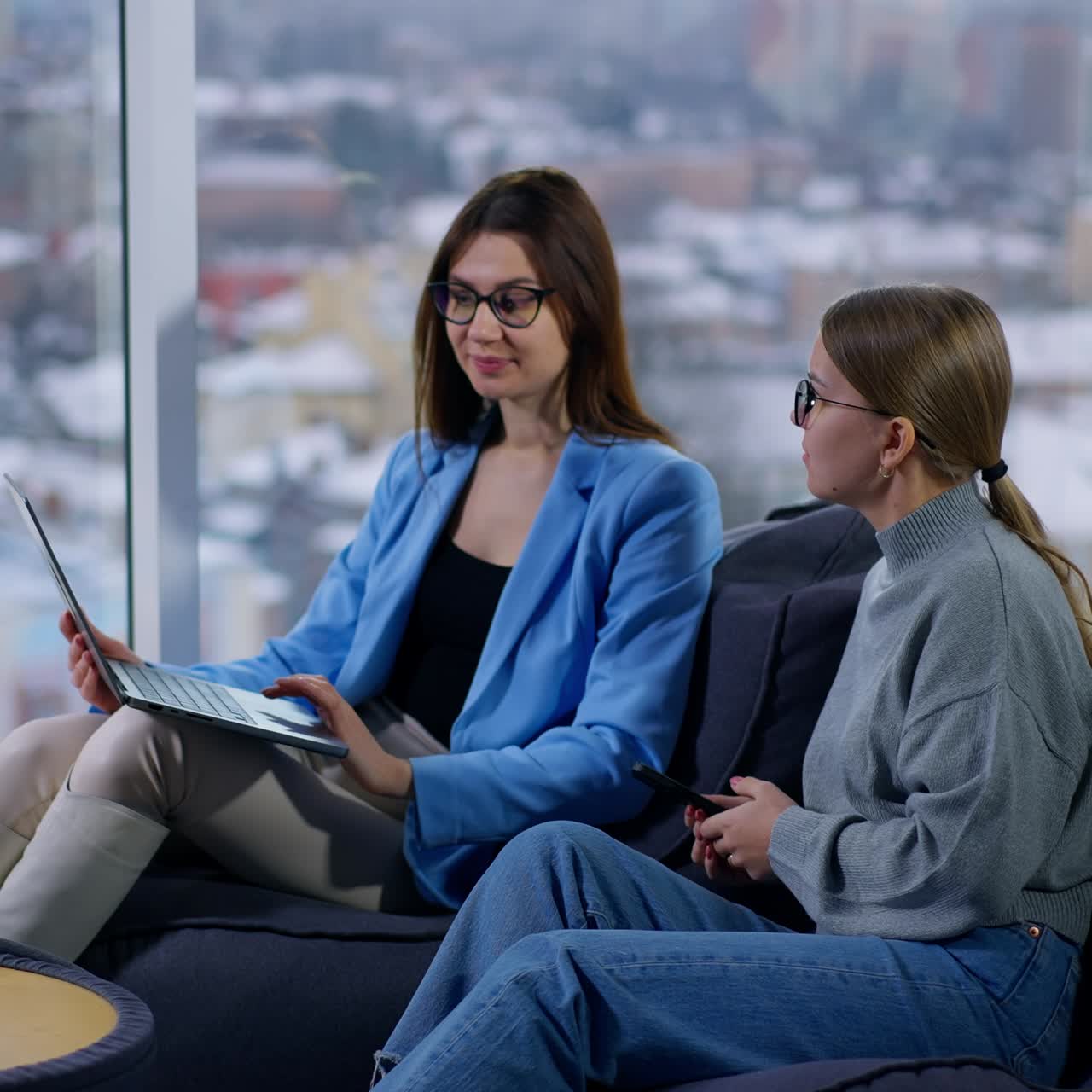 Due colleghe caucasiche parlano di questioni lavorative. Le donne usano un laptop sedute su pouf a sacco. Finestra panoramica con paesaggio urbano sfocato sullo sfondo