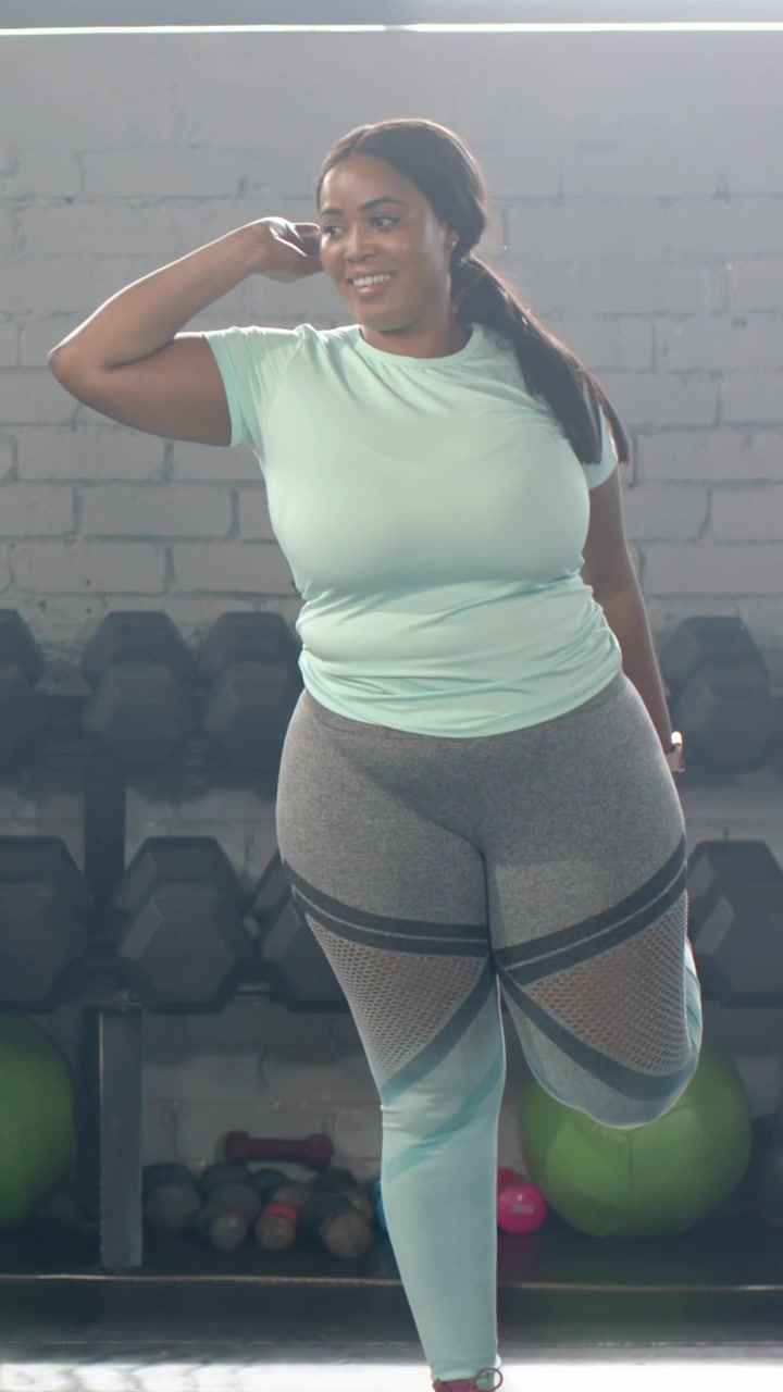 Video verticale: Donna con top color menta che solleva la gamba destra e mantiene lo stretching per il riscaldamento in palestra con i pesi.