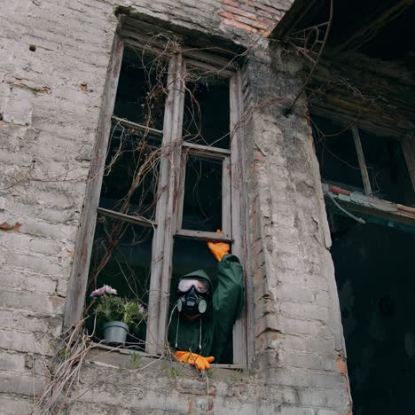 Ecologist wearing respirator alone in empty house. Man in safety suit and mask looking out of the window without glass from abandoned building. Chemical attack