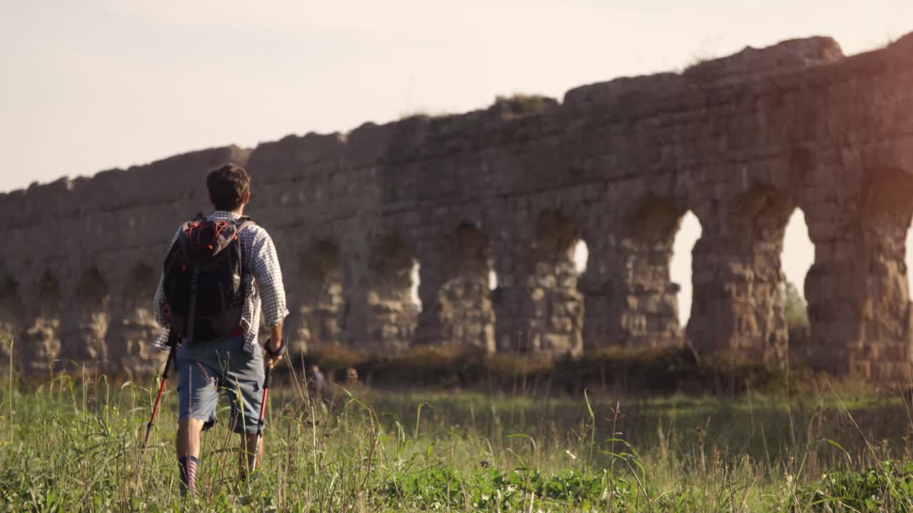joven mochilero camina descalzo con palos en la hierba frente a los arcos del acueducto romano en el parco degli aquedotti ruinas del parque en roma al atardecer en cámara lenta