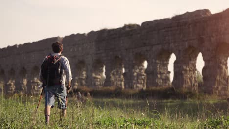 young man backpacker walks barefeet with sticks on grass in front of roman aqueduct arches in parco degli acquedotti park ruins in rome at sunset slow motion