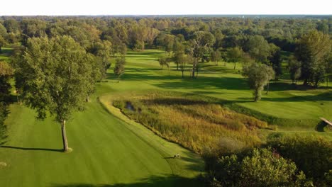 Aerial-establishing-drone-shot-flying-upfront-about-trees-showing-golf-course-with-pond-during-sunny-summer-day