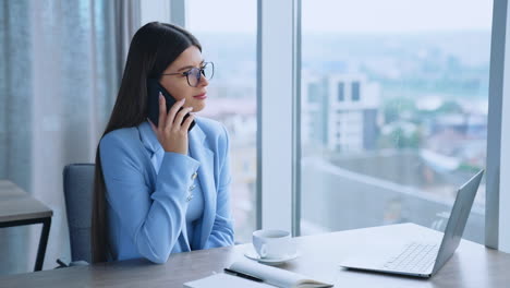 beautiful brunette lady sitting at desk in the office having phone conversation. woman in glasses talking with clients, discussing and solving problems. close up.