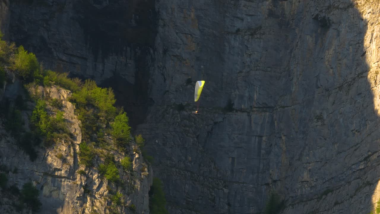A person is paragliding with giant rocks in the background. The person appears extremely small. He is practicing his sport on a beautiful spring day, and the trees on the rocks are illuminated.