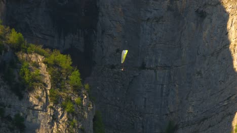 a person is paragliding with giant rocks in the background. the person appears extremely small. he is practicing his sport on a beautiful spring day, and the trees on the rocks are illuminated.