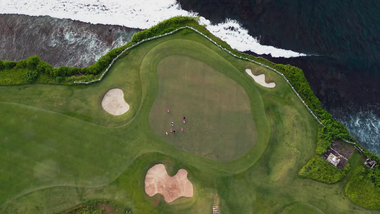 Aerial drone shot of coastal golf course showing golfers on the green field with open ocean reef, below white sand shoreline and tall limestone cliffs in clear daytime