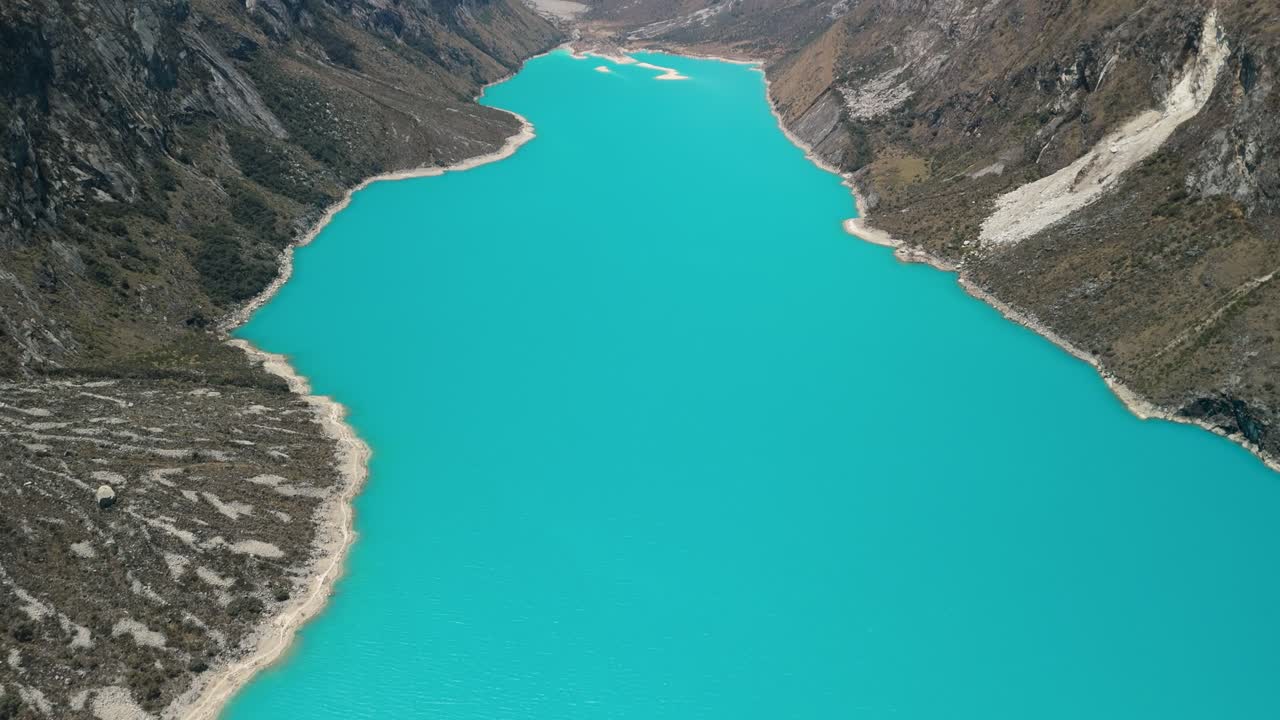 A breathtaking high-altitude aerial shot tilts up from the vibrant turquoise water of Laguna Parón to reveal the spectacular snow-capped peaks of the Cordillera Blanca in the Peruvian Andes
