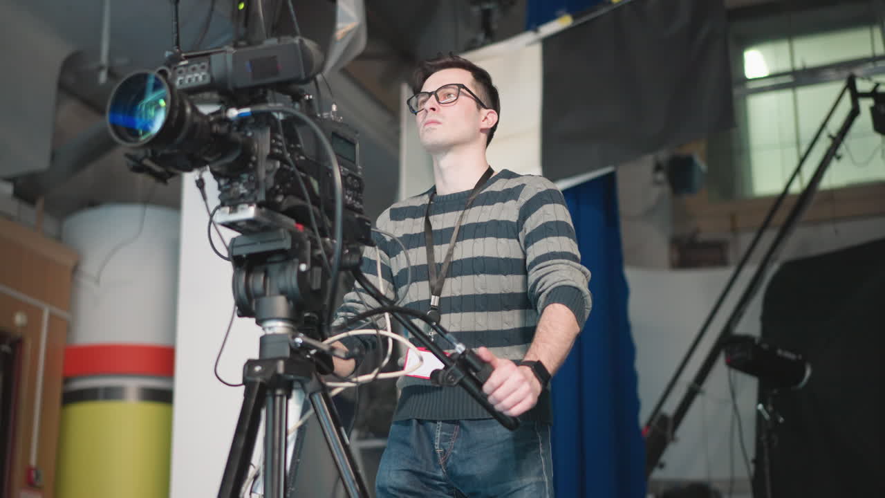 Camera operator adjusting equipment in TV studio, focusing on camera while preparing for filming. Studio setup with crew member operating high-end broadcast camera on tripod