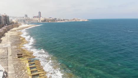 Sliema-rocky-seascape-blocked-by-Promenade-and-city-skyline,-in-Malta---Revealing-Pan-aerial