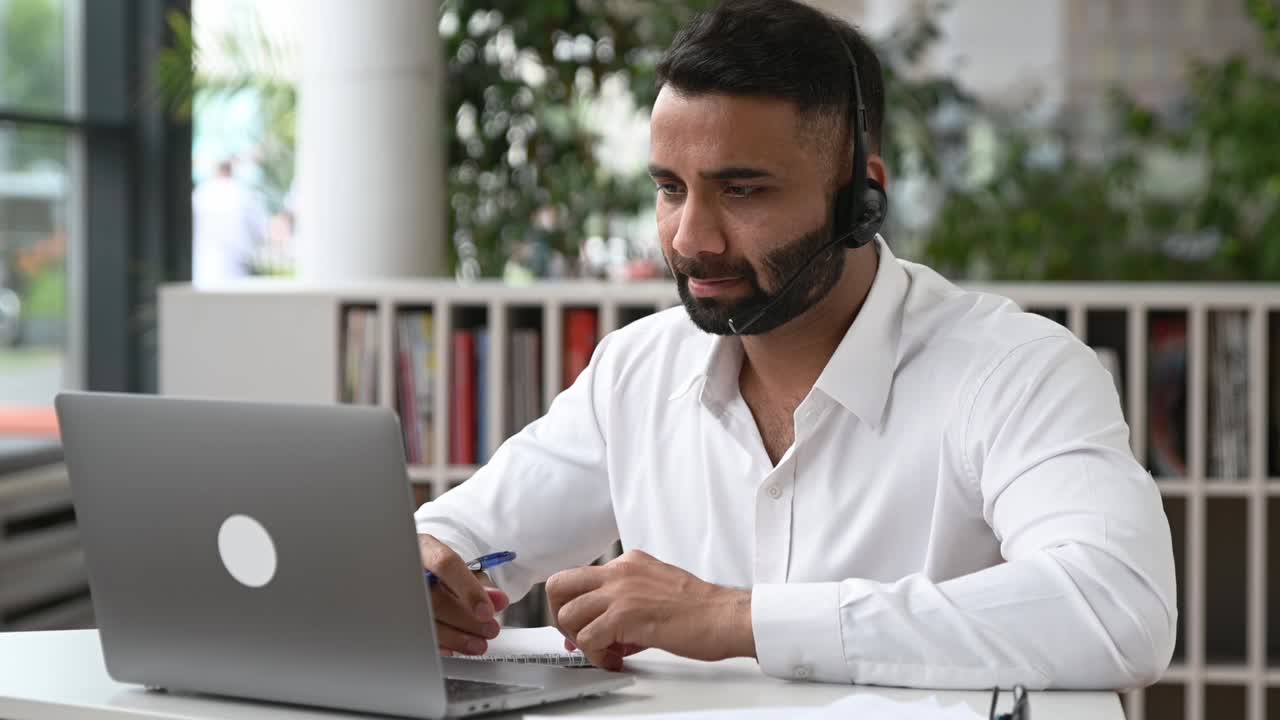 Uomo indiano intelligente, determinato e attraente con cuffie, manager, operatore di supporto, consulente online, dirigente, seduto al laptop in un ufficio moderno, che ascolta, registra una richiesta e svolge una consulenza online
