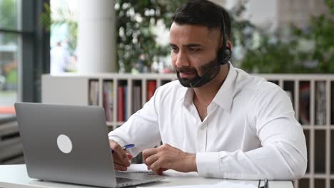 smart purposeful attractive indian man with headset, manager, support worker, online consultant, executive sitting at laptop in modern office, listening, recording appeal, doing online consultation