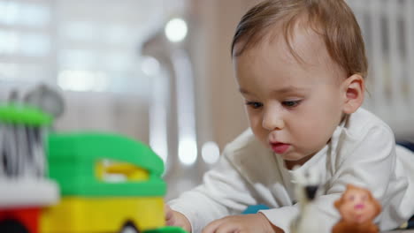 cute calm baby boy in white shirt lies on belly on the floor. sweet little toddler is interested with a toy car in front of him. close up. blurred backdrop.
