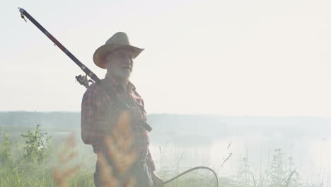 Side-view-of-old-man-in-a-hat-standing-on-the-shore-of-a-lake-with-a-fishing-rod-on-a-cloudy-morning