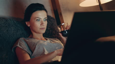 mujer usando computadora tarde en la habitación. chica trabajando en un cuaderno con un vaso de vino.