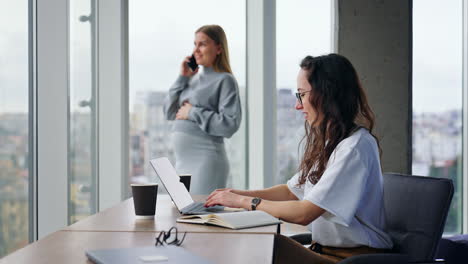 young mid-aged ladies working in the modern spacious office. brunette works at her laptop. pregnant colleague speaks on the phone standing near the window.