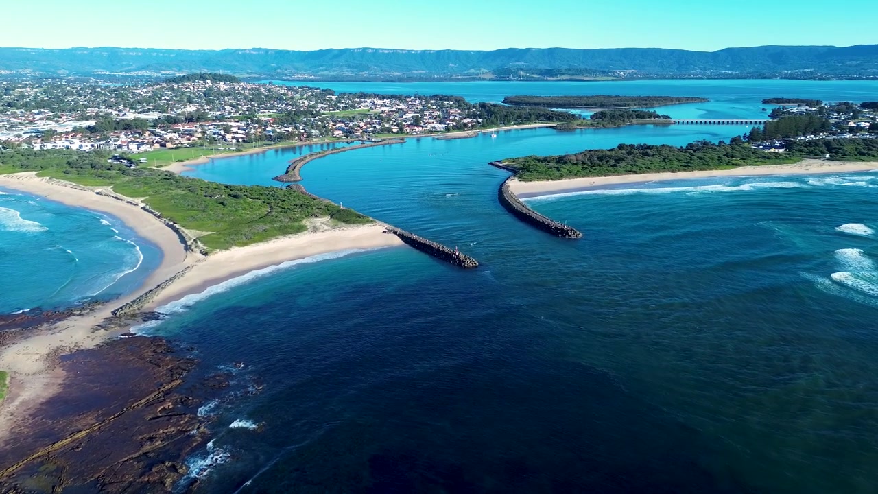 Paesaggio aereo con drone della diga frangiflutti oceanica di Windang Passage presso la rocciosa spiaggia di Warilla, con una città costiera suburbana e l'insenatura del Lago Illawarra sullo sfondo, vicino a Wollongong, costa meridionale dell'Australia, turismo.