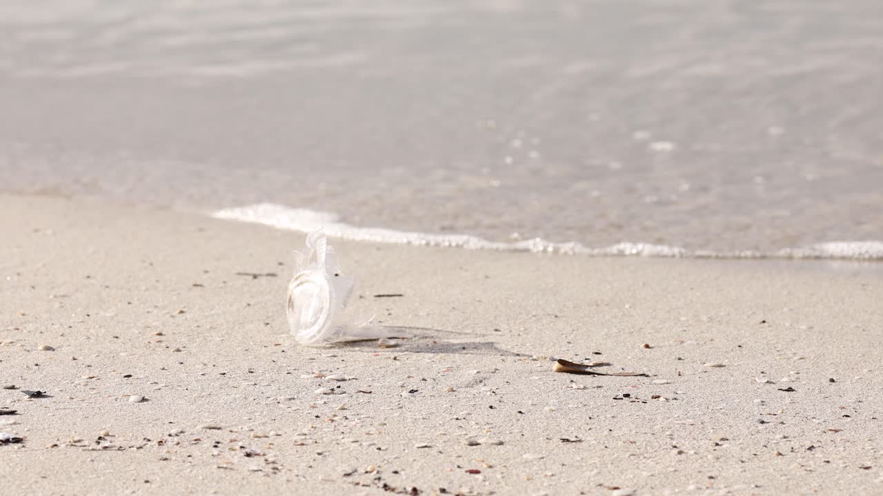 모래 해변에 놓인 버려진 플라스틱 컵, 배경에 파도, 환경 문제
