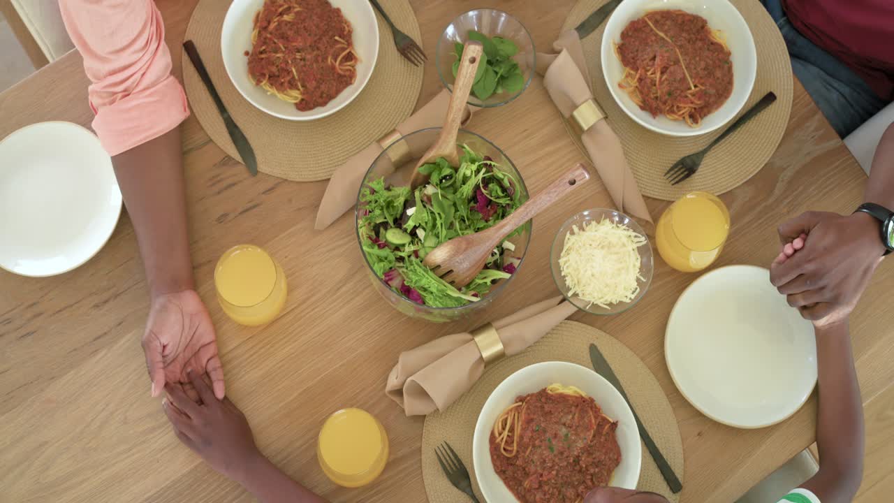 Famiglia afroamericana che si tiene per mano a tavola mentre la mamma si protende verso il bambino, lasciandolo andare, per iniziare a mangiare gli spaghetti.