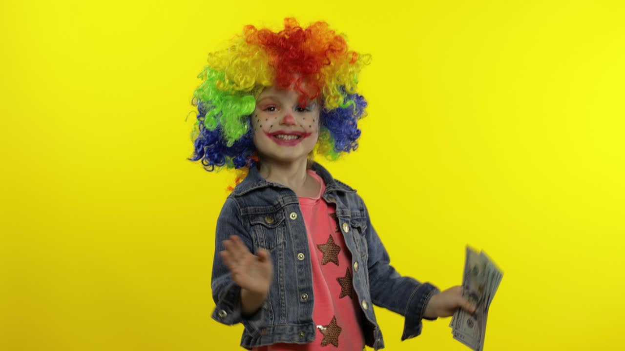 niña payaso en peluca colorida bailando con dinero dólares billetes en efectivo. tonto alrededor, sonriendo
