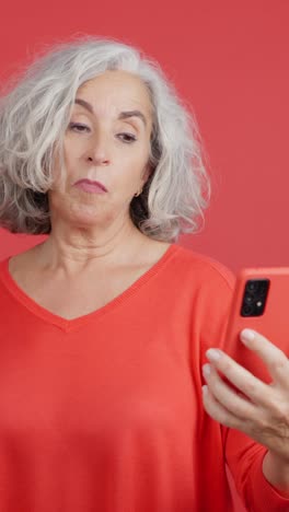 Confused Senior Woman Looking at Smartphone on Red Background