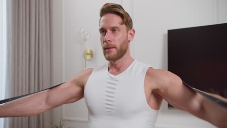 a confident blond man with an athletic build and stubble in a white t-shirt makes swings with his arms and does morning exercises using a special elastic band in a modern apartment in the morning at home