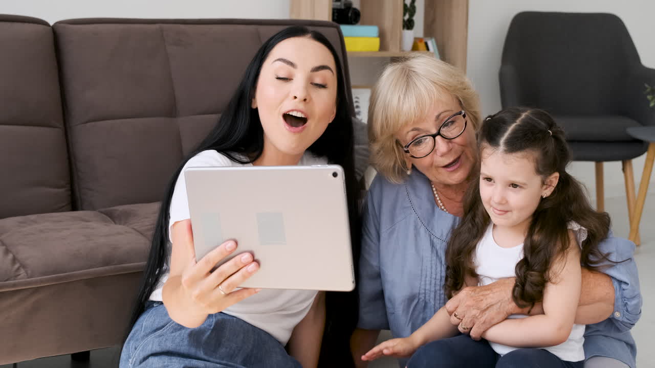 家の床に座っているタブレットを介してビデオ通話で話している間、彼女のお母さんとおばあちゃんの挨拶を持つ愛らしい少女