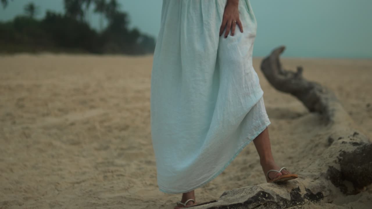 Persona caminando por la playa al atardecer, descalza con un vestido blanco elegantemente drapeado, tocando madera flotante