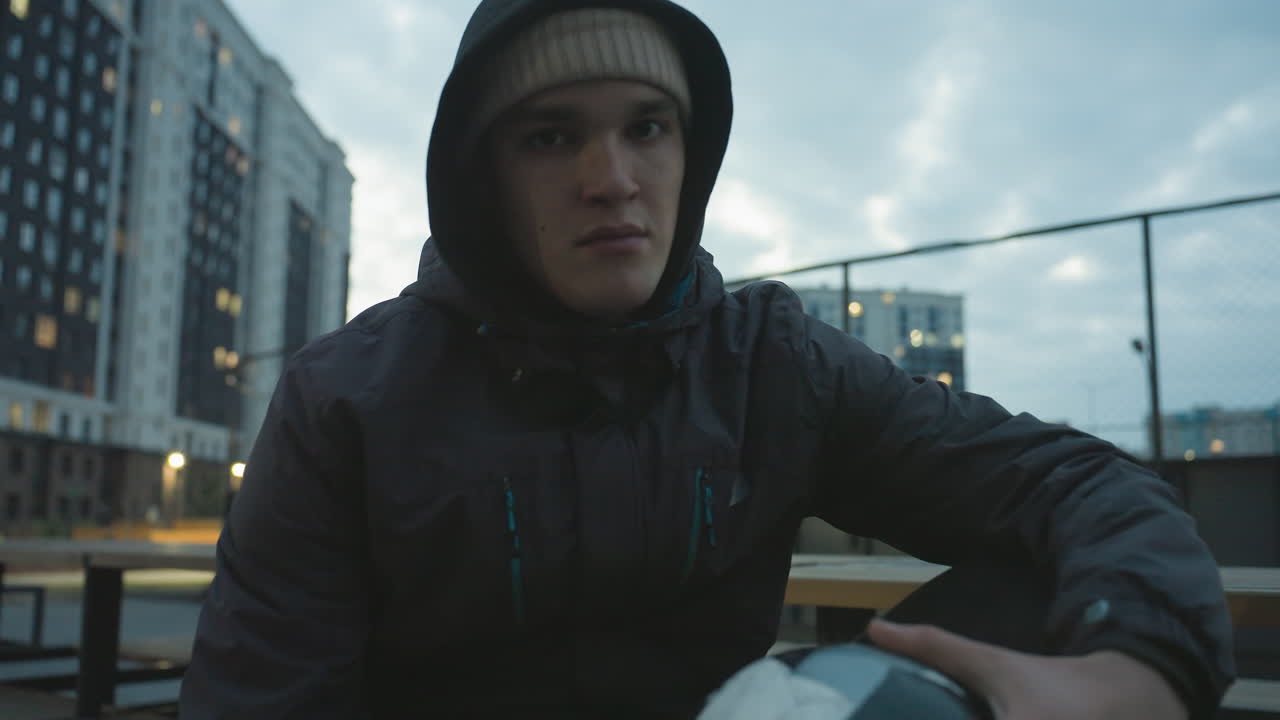 Man in hoodie sitting on bench gripping soccer ball rotating it with intense focus during urban training session, surrounded by modern residential buildings and evening city lights