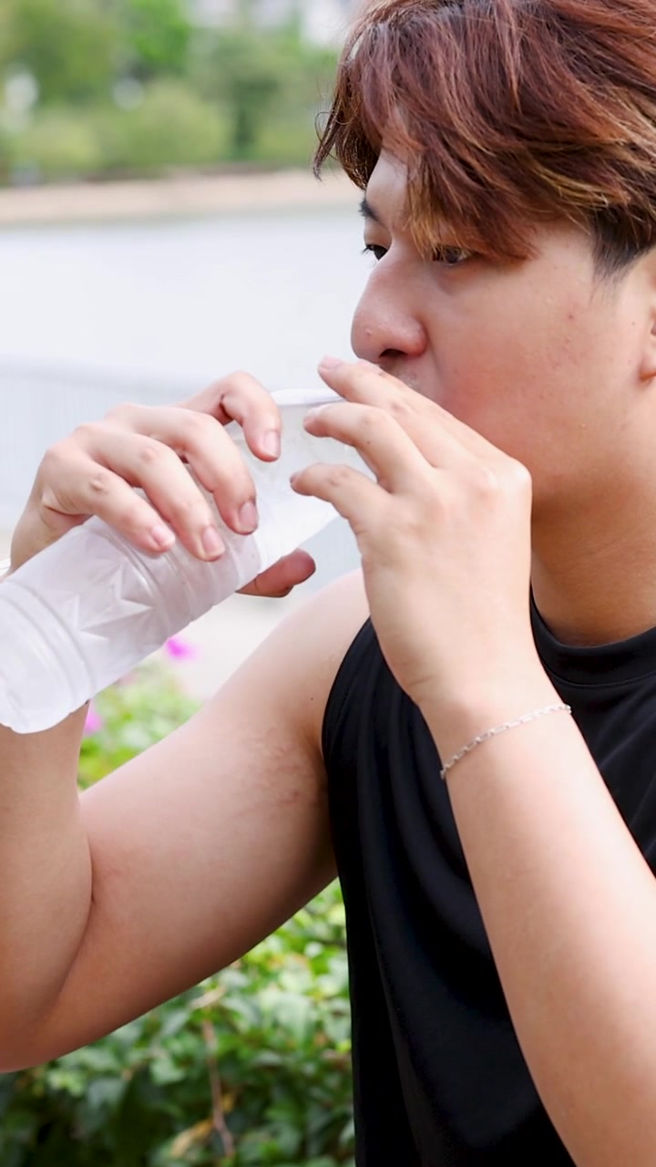 Seorang pria muda Thailand mengenakan kaus tanpa lengan hitam minum air kemasan di luar ruangan.