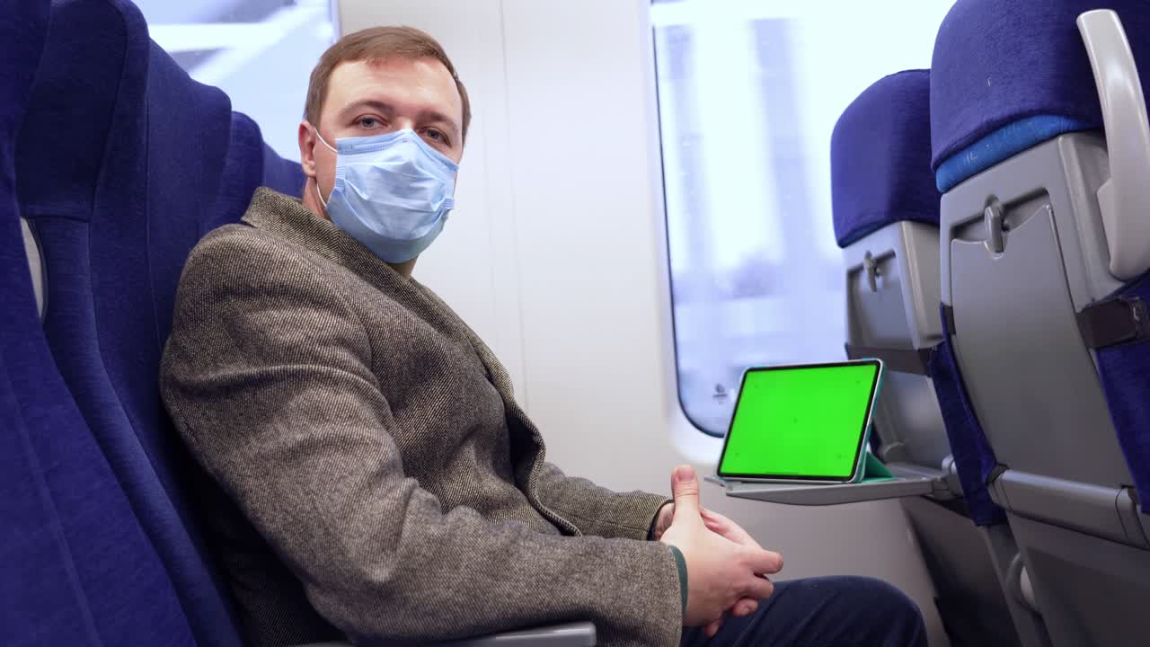 코트와 의료 마스크를 입은 밀레니엄 남자가 기차를 타고 카메라를 바라보고, 그의 앞에 접이식 테이블에 크로마 키 녹색 화면으로 된 태블릿 컴퓨터가 있습니다.