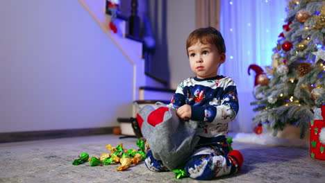 adorable child in warm winter pajama plays near the christmas tree. cute baby boy getting the sweets from stocking.