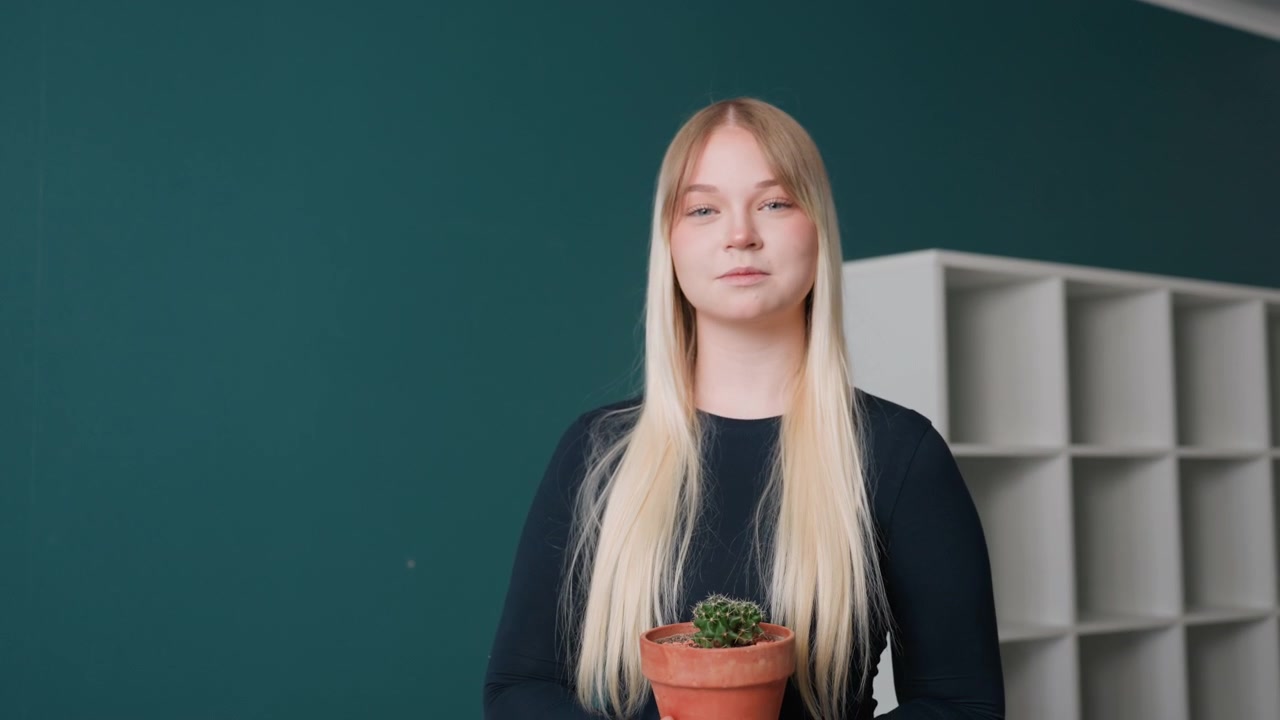 Weiße Frau hält Pflanze, selbstbewusste Haltung, neue Mieterin posiert in inszeniertem Wohnbereich mit petrolfarbener Wand und offenen Regalen, langes blondes Haar, Terrakotta-Topf, direkter Blick, professionelle Ausstrahlung, leichtes Lächeln