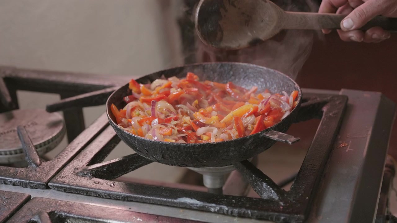 Pimientos y cebollas salteándose en sartén de hierro fundido sobre quemador de gas, con vapor, cocción a fuego alto y preparación culinaria artesanal, toma estática de cocina en cámara lenta