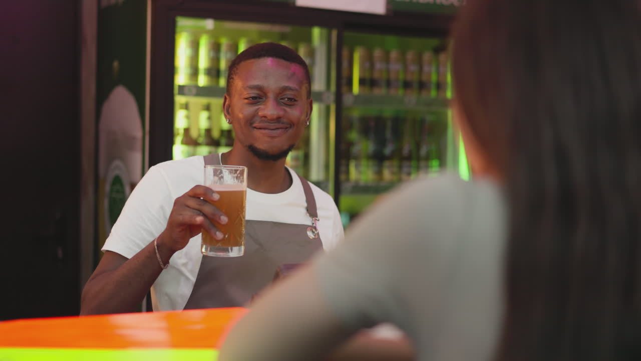 Flirting barman offers beer to woman at counter. Cheerful barkeeper gives glass of lager to pensive girl guest in night club. Black man treats lady