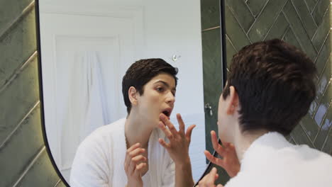 happy caucasian woman looking in mirror, touching face and hair and smiling in bathroom