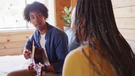 Happy-african-american-couple-playing-guitar,-sitting-on-sofa-in-log-cabin,-slow-motion