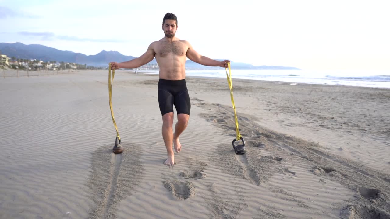 metabolic workout on the beach with muscular man performing back and rubber variant with kettlebell at sunrise