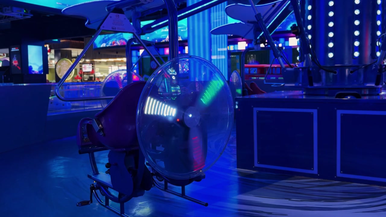 Un'inquadratura statica di una giostra meccanica vuota all'interno di un centro di intrattenimento familiare buio. Il sedile a pedali presenta una ruota che gira illuminata da luci al neon blu brillante e LED