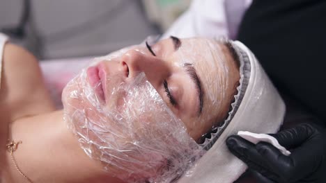 Professional-cosmetologist-is-cleaning-woman's-face-from-special-treatment-using-cotton-sponge.-Young-woman-is-lying-on-the